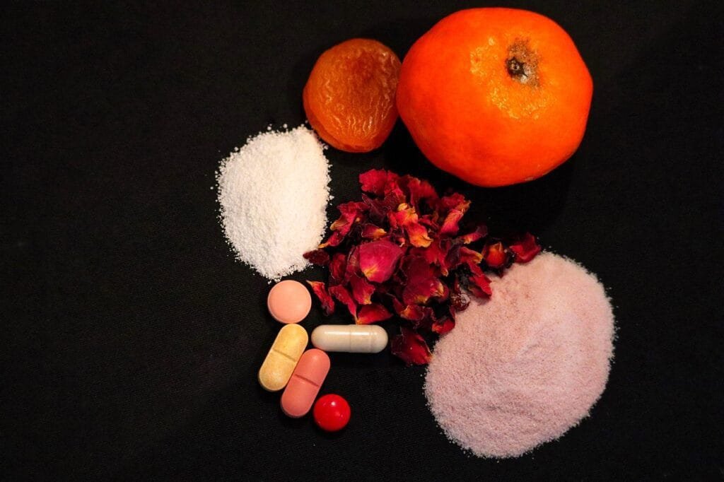 Eine Vielzahl von Nahrungsergänzungsmitteln, darunter Tabletten, Kapseln und rosa und weiße Pulver, arrangiert mit einer frischen Orange und getrockneten Früchten