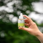 Nahaufnahme einer Person, die eine durchsichtige 30-ml-Flasche Vita Elixia Bioactive Prebiotics Jelly Serum vor einem weichen, verschwommenen Hintergrund mit üppigen grünen Bäumen hält.