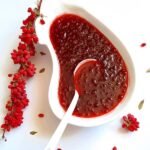 Eine weiße, nierenförmige Schale, gefüllt mit leuchtend roter Bio-Berberitzenmarmelade, abgebildet mit einem Löffel und einem frischen Zweig leuchtend roter Berberitzen an der Seite